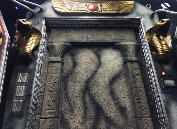 In-game: A sealed stone doorway to an ancient Egyptian tomb flanked by king cobra statues and asurrounded by strange technology.