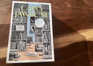 The box art for Cain's Jawbone depicts a library with a deadman on the floor and a person in the shadows outside of the window.