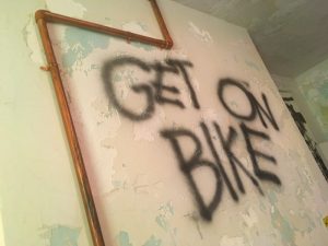 In-game: "GET ON BIKE" is spray painted on an old plaster wall.