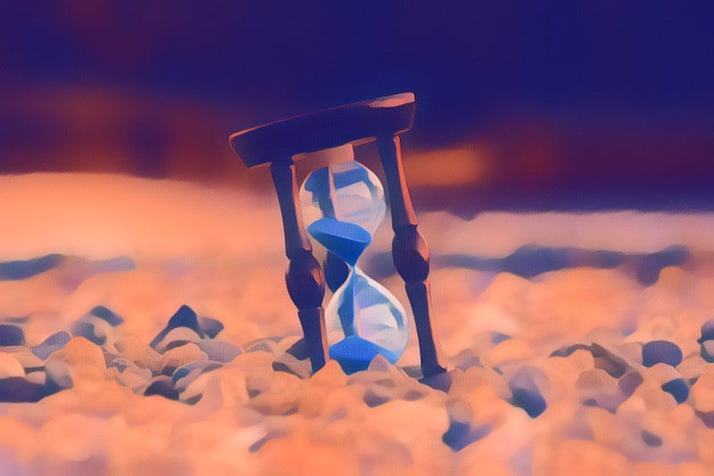An hourglass with blue sand sitting atop pebbles.