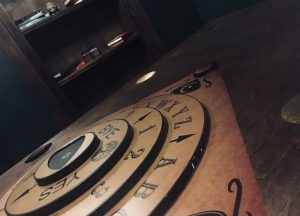 In-game: Closeup of a twistable Ouiji board with a bookcase in the background.