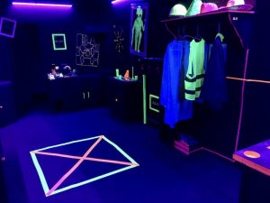 In-game: wide shot of Darklight Disco. There are an assortment of costumes hanging from the wall, and a glowing square on the floor.