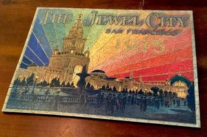 Image of an intricate wooden jugsaw puzzle depicting art from the San Francisco World's Fair. "The Jewel City" shows a tower with lights matching teh colors of the rainbow emerging from it.