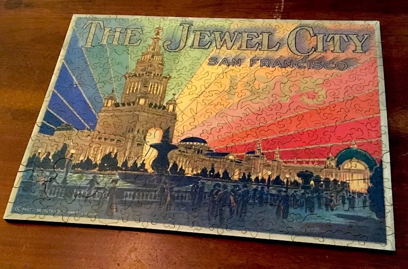 Image of an intricate wooden jugsaw puzzle depicting art from the San Francisco World's Fair. "The Jewel City" shows a tower with lights matching teh colors of the rainbow emerging from it.