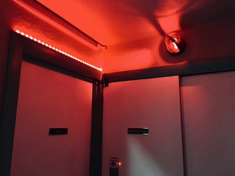 In-game: a small dim room lit by a red alarm light.
