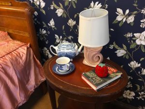 In-game: closeup of a nightstand with a book, a lamp, a tea pot and a tea cup.