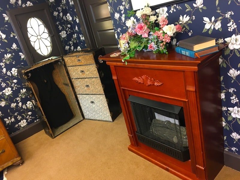 In-game: an open trunk with a dress hanging inside beside a fireplace.