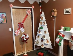In-game: A white christmas tree shelf covered in gingerbread cookie ornaments beside a fireplace decorated with stockings.