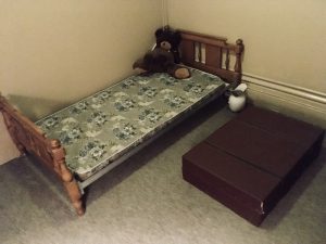 In-game: A ratty bed with a large stuffed animal on it. Beside the bed on the floor is a massive briefcase.