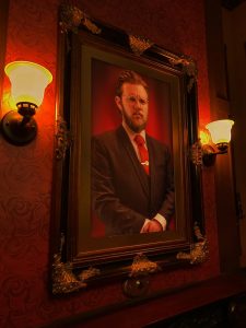 A portrait of a stern-looking bespectacled man in a suit, lit by sconces.