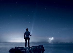 A lone person standing on a island in the middle of the water holding a flighlight in the darkness.
