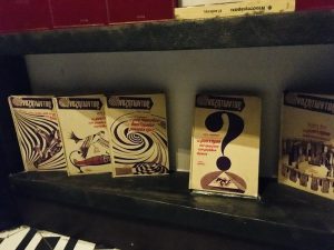 In-game: Closeup of a collection of books written in Greek. The Center book has a large question mark on the cover.