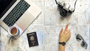 A hand gesturing to a place on a map. Atop the map is a camera, passport, sunglasses, laptop, and a cup of coffee.