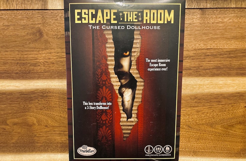 The Cursed Dollhouse box art depicting a creepy doll peering through the cracked wall of a dollhouse.