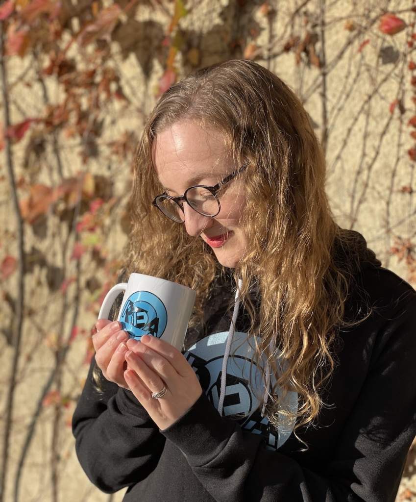 Lisa looking lovingly into a REA coffee mug while wearing a REA hoodie on an autumn day.