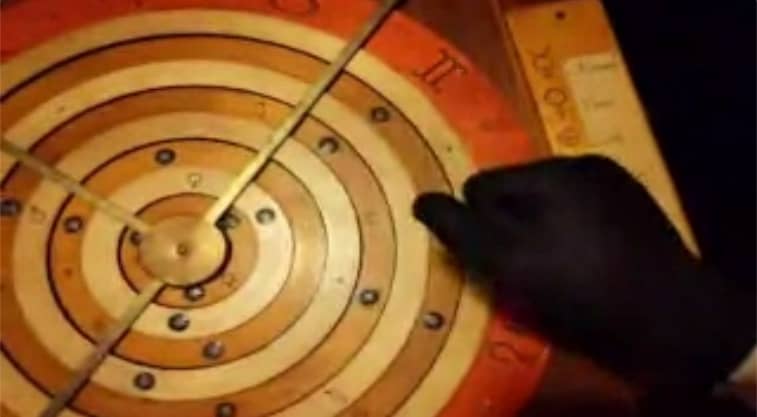 A black-gloved hand manipulating an astrology puzzle. The text on the puzzle is difficult to read.