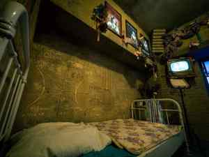 bed with metal frame in a dingy room with scribbles on the wall and photos above.