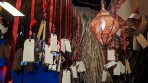 Closeup of a large object with many red ribbons and tags hanging from it, illuminated by an ornate lantern.