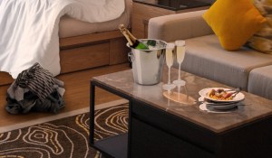 Image of a hotel room the morning after a fun night. Champagne, and room service dishes sit on a table beside an unmade bed with clothes on the floor.