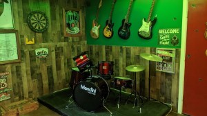 A drum kit on a small riser in a dive bar. The wall beyond it has a dart board, beer ads, and 4 electric guitars hanging.
