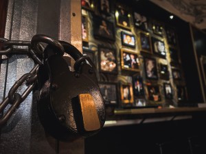 Close up on an old lever lock, securing a magical cabinet. Beyond it is a wall covered in photos of famous magicians.