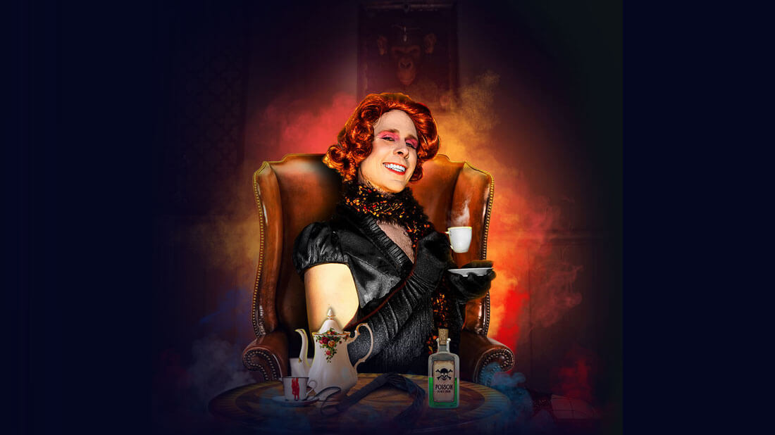 a man in drag, wearing a red wig sitting in an easy chair holding a teacup. on the coffee table in front is a tea pot and a bottle of poison.