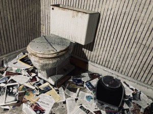 A gross toilet locked shut, the floor below is covered in torn up paper.