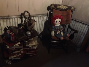 A large redheaded doll sitting on a rocking chair, beside it is a stack of old books and empty beer cans.