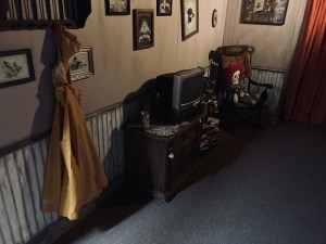 An old, rundown livingroom with a yellow rain coat hanging from a hook, a doll on a rocking chair, and an old TV with empty beer cans beside it.