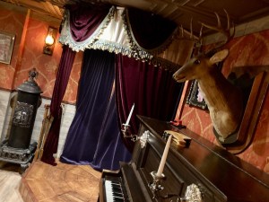 A buck taxidermy mounted above a piano beside a stage in a western saloon.