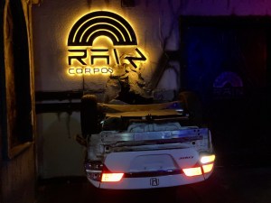 A large, illuminated, and broken Rain Corporation logo mounted to a wall, with an upside dow Honda Civic crashed through the wall.