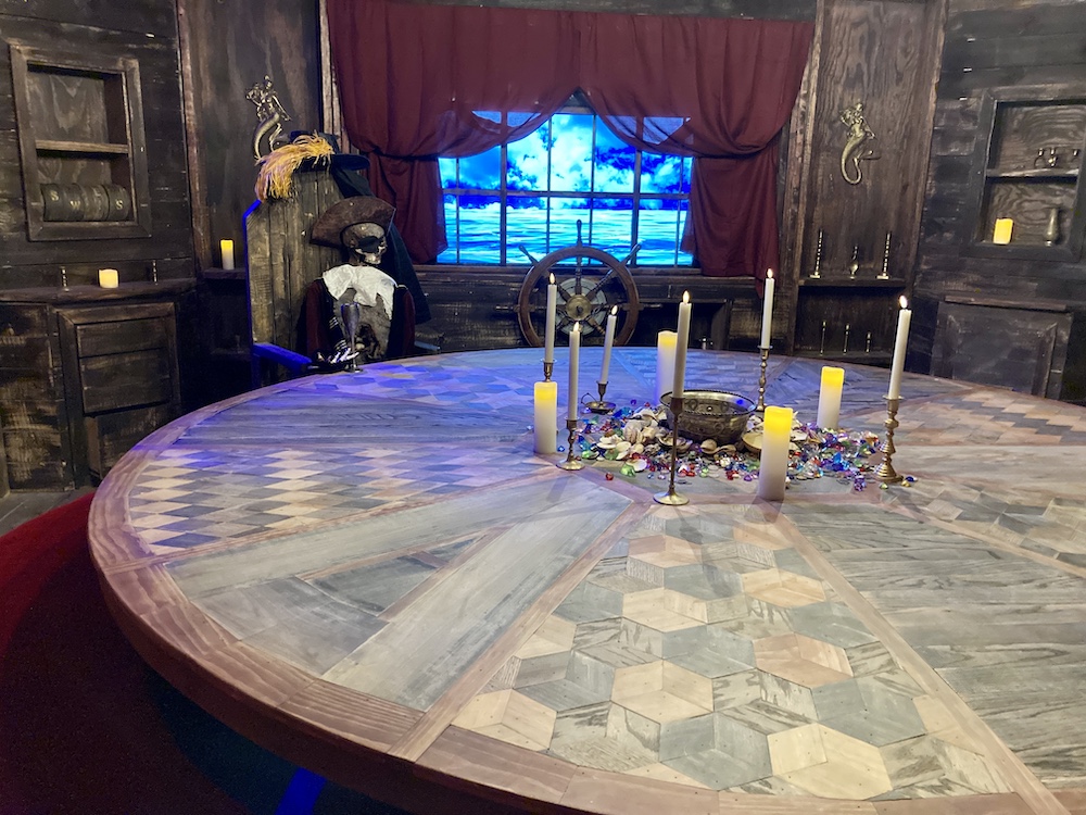 A large, intricately designed circular wooden table on the other end is the skeleton of a pirate captain in a chair beside a ships wheel overlooking the sea.