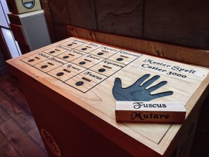 A large wooden device named teh "Master Spell Caster 3000" has a hand print on it beside a grid of magical words.