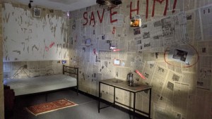 A room with walls covered in newspaper, in big red letters are the words, "Save him!" painted on the walls.