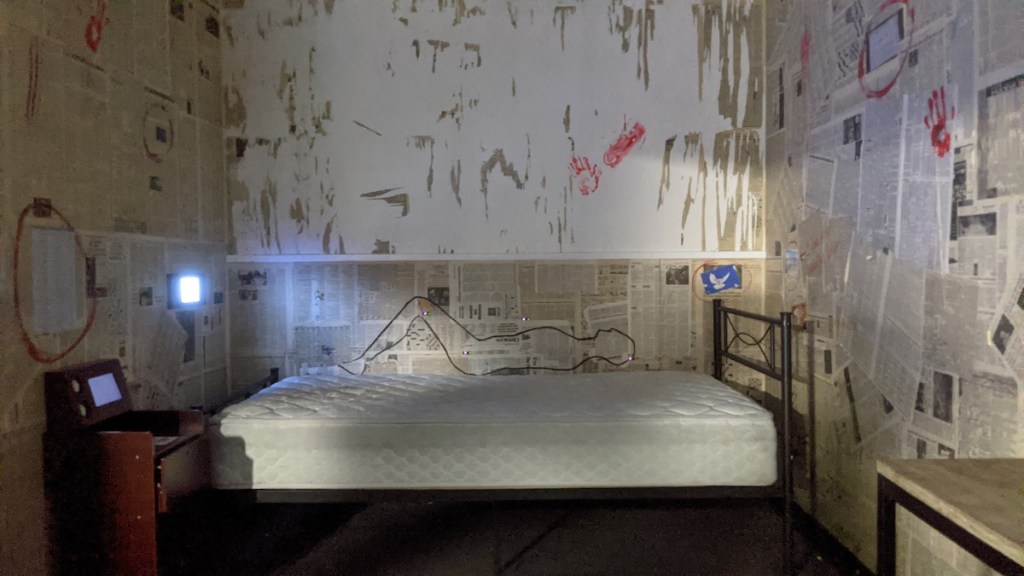 A mattress in the middle of a room covered in newspaper, just above the mattress is a drawing of a man.