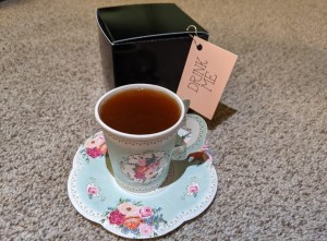 A tea cup filled with tea on a saucer beside a black box labeled, "drink me."