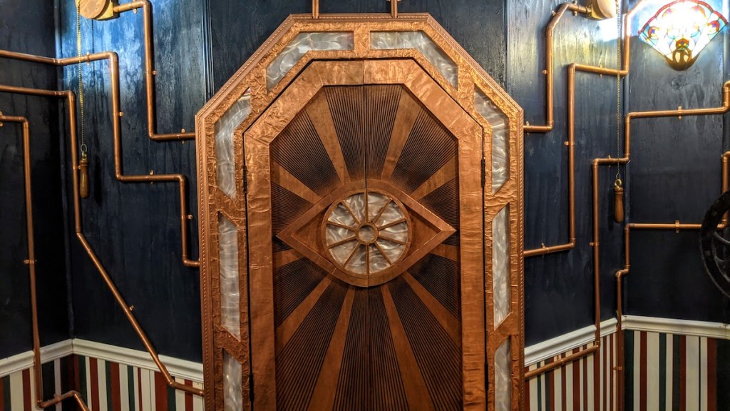 An ornate bronze door with an eye. Copper pipes emanate from the door.