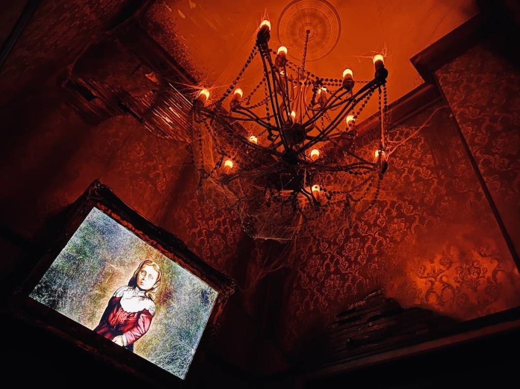 Upward view of a room in a haunted house. Everything is damaged and covered in cobwebs. Below is a glowing portrait of a young girl.