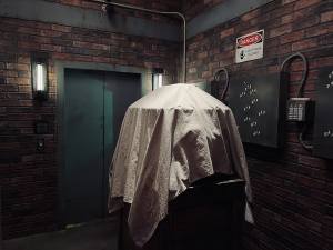 A box covered in a white sheet in a warehouse. Beyond it is an elevator door and electrical panels.