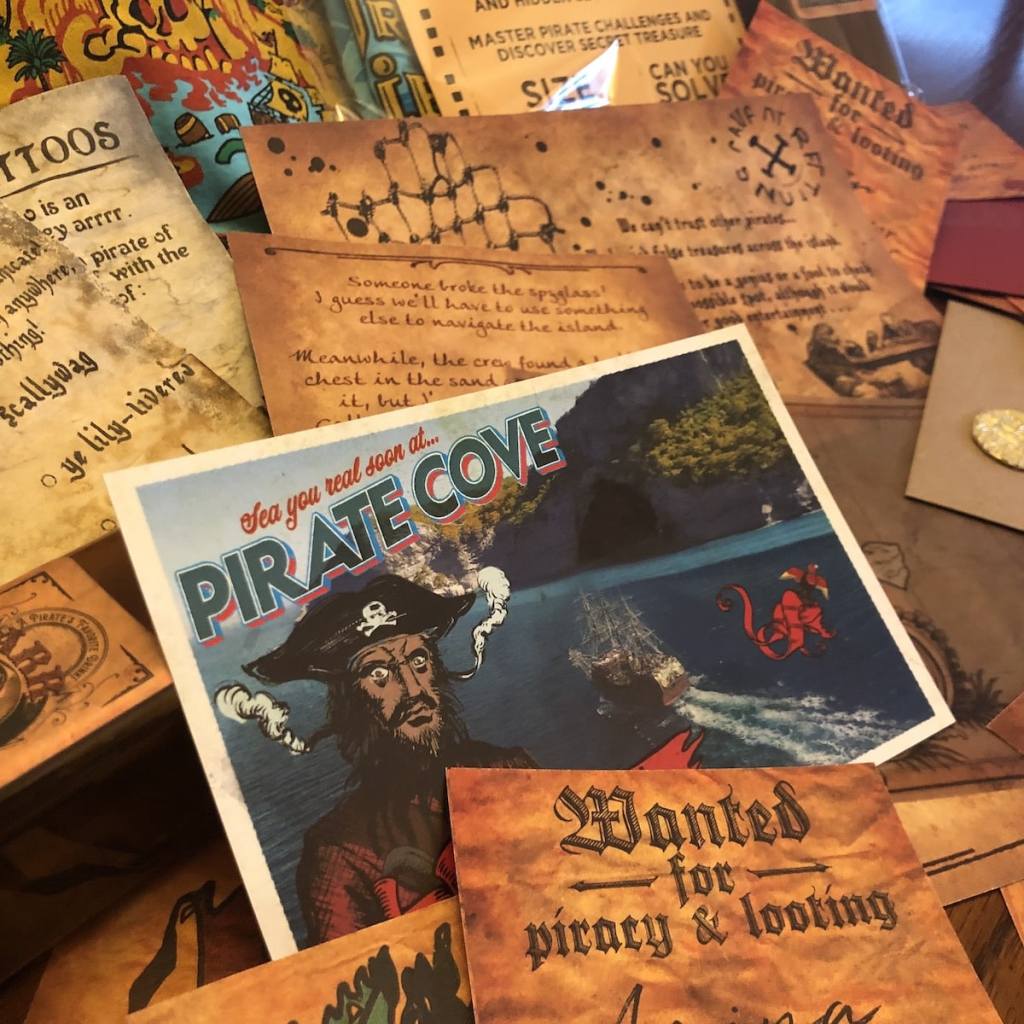 An assortment of aged paper items from the game, in the middle is a post card with a pirate on it that says, "Sea you real soon at... Pirate Cove."