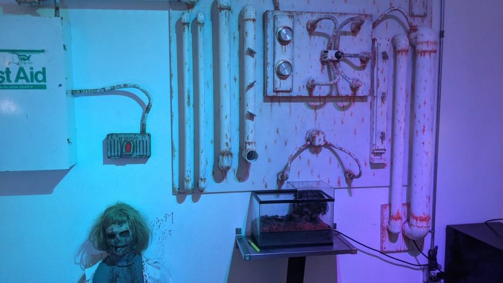 A wall covered in corroded pipes a zombie leans against the wall.