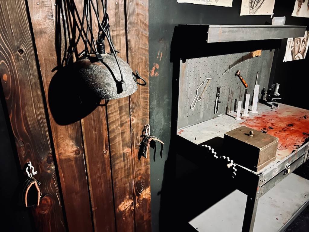 A helmet connected with wires to a strange looking torture bed, beside a workbench.