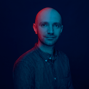 Man with shaved head in a button down denim shirt with a blue background and a red light shining on the left half of his face.