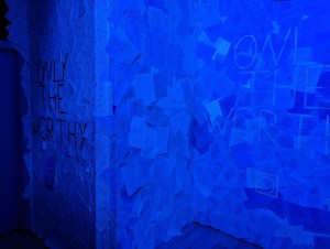 A wall covered in scraps of paper, the words "ONLY THE WORTHY" are written in black ink and again in UV ink.