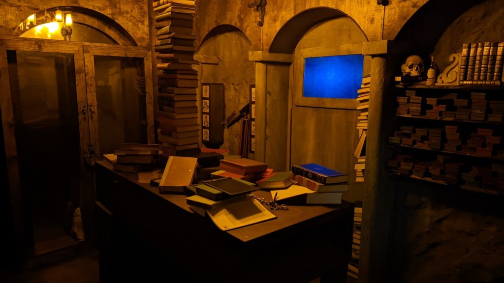 A wizard's library with a desk covered in books. In the middle of the desk is a very tall stack of books.