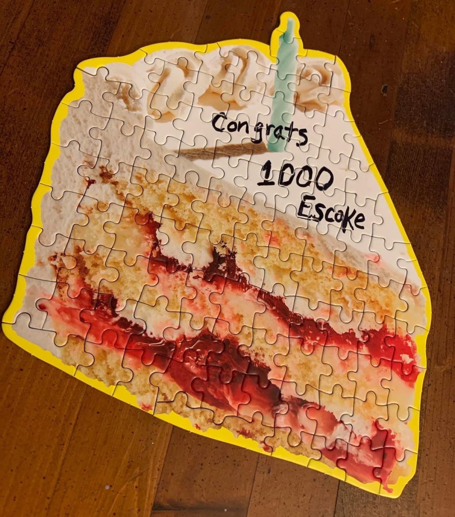A jigsaw puzzle of a slice of cake with a candle. Handwritten on the top are the words, "Congrats 1000 Escake"