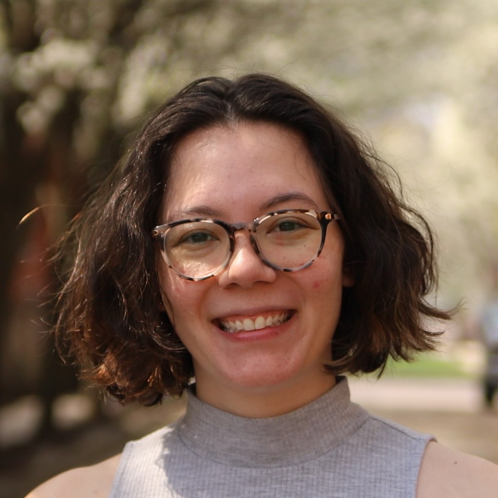 woman of mixed asian descent with short wavy hair in glasses smiling, wearing a grey sleeveless mock turtleneck.