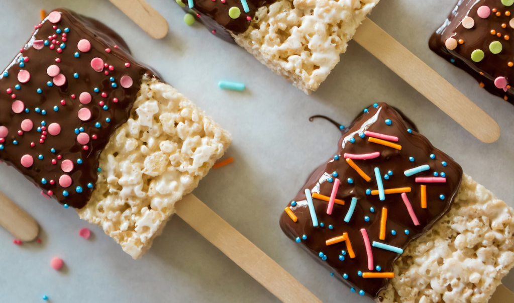 Rice crispy treats covered in chocolate and sprinkles on popsicle sticks.