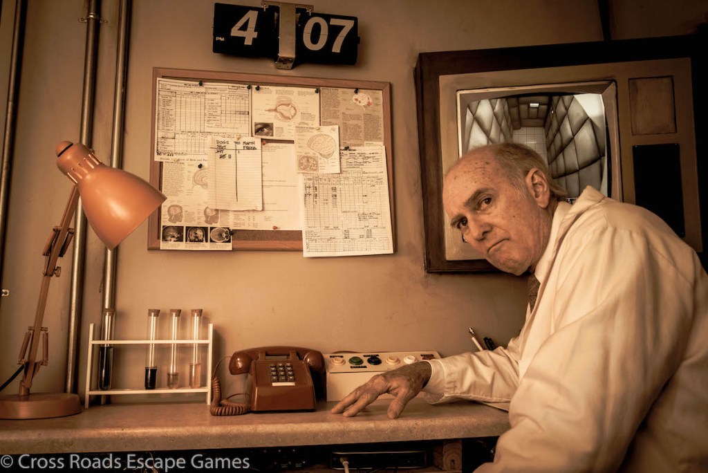 Ominous-looking Dr. Griffen at a control station with a phone and an assortment of buttons and papers in front of him.