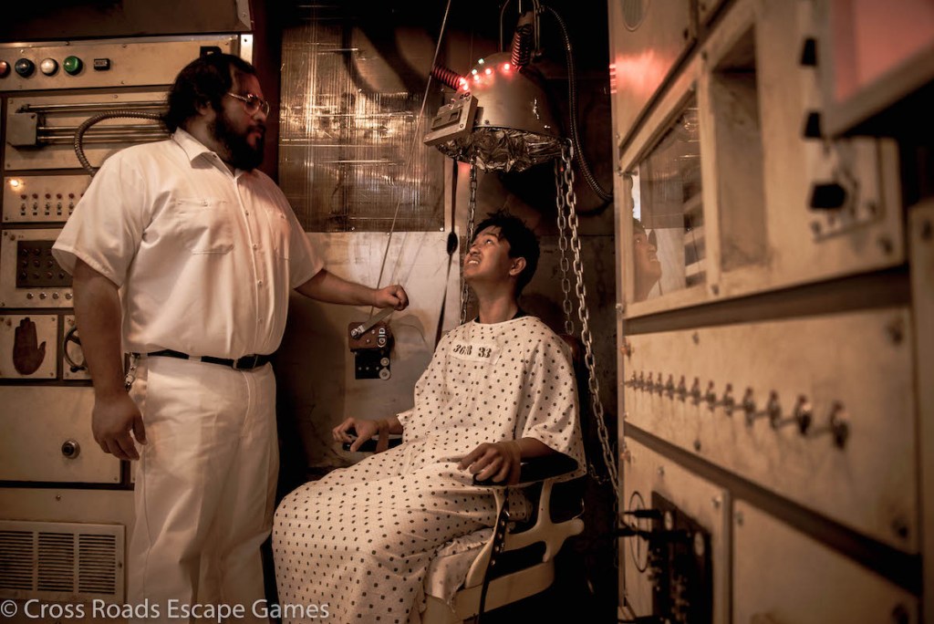 An orderly lowering a strange helmet on a patient strapped to a chair.